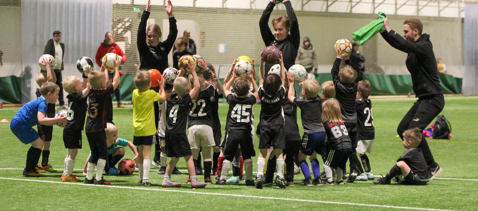 Ryhmä pieniä lapsia leikkii kolmen ohjaajansa johdolla TPS:n jalkapallokoulussa vanhempien seuratessa sivusta.